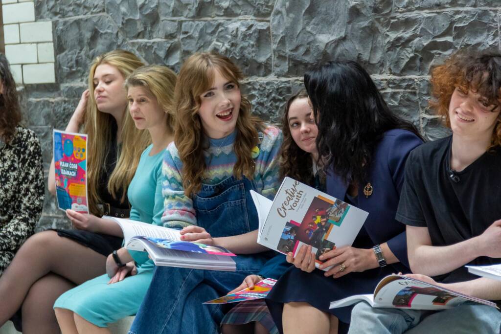 Young people sit talking and smiling in front of a stone wall with Creative Youth pamphlets