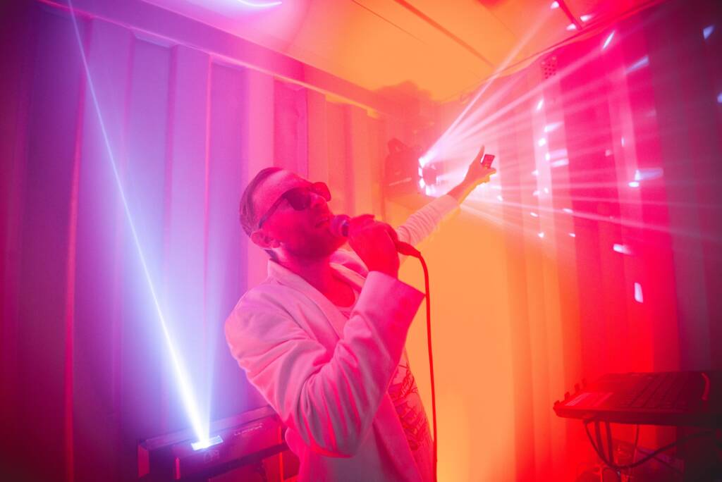 A performer sings into a microphone and points upwards. He wears a white suit jacket and sunglasses. He is surrounded by strobe lights in a room lit in pink yellow and purple