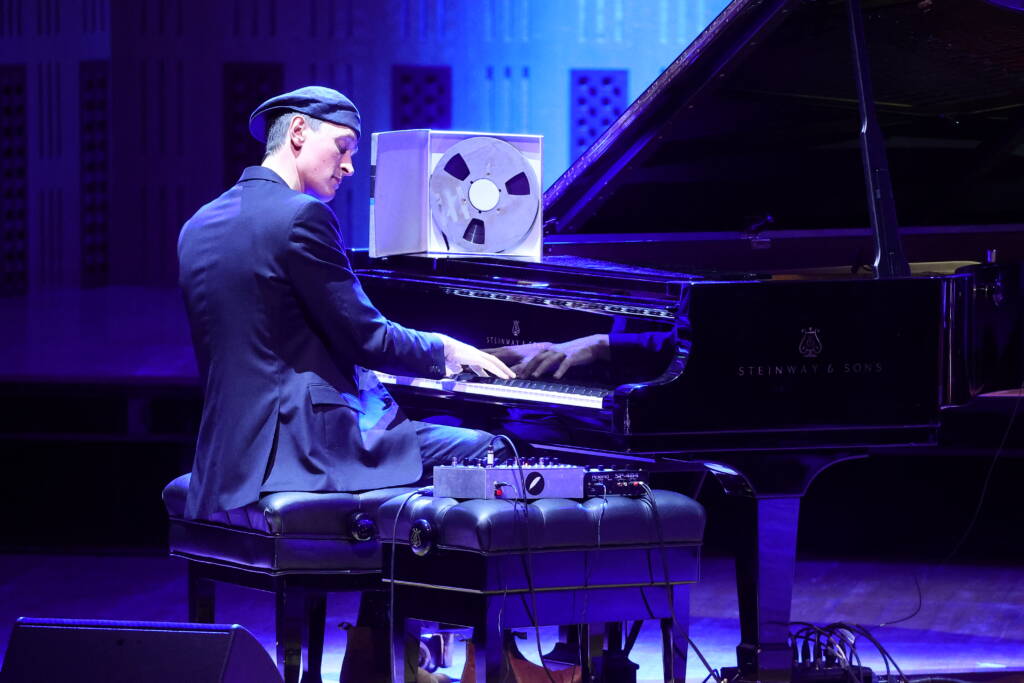 Erland Cooper sits playing the piano. He wears a suit and a backwards flat cap. His eyes are closed as he plays, to his right is a musical switch baord and in front of him is a fil;m reel. He is lit in blue