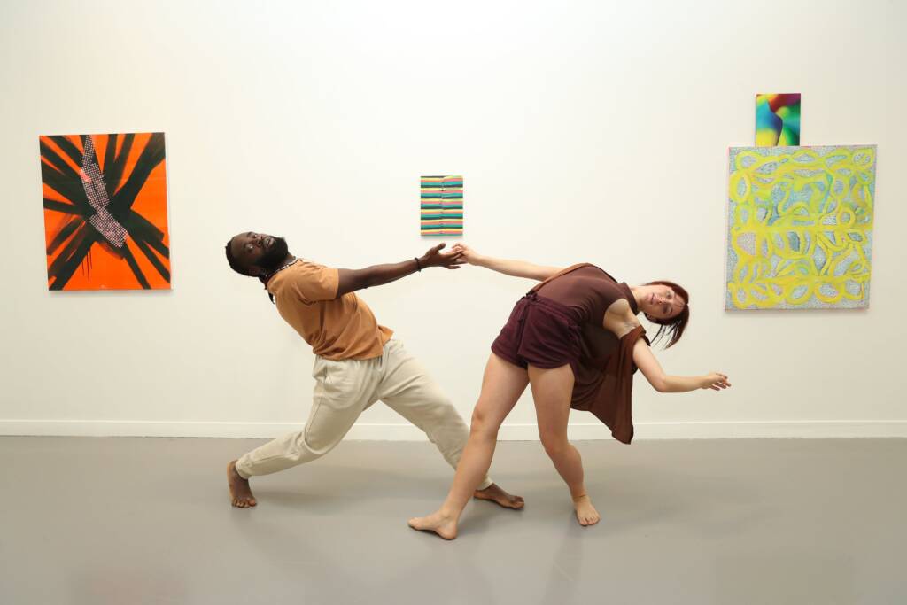 Two dancers are caught in motion in front of a gallery wall. They bend away from each other as their outstretched hands almost touch.