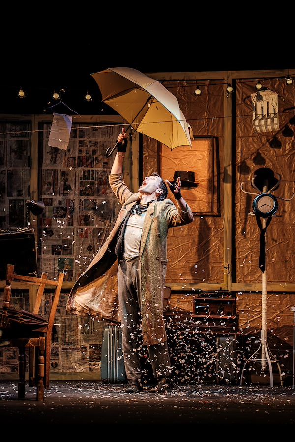 A performer stands on stage holding an umbrella and dramatically raises their arms to the sky. Confetti swirls around them.
