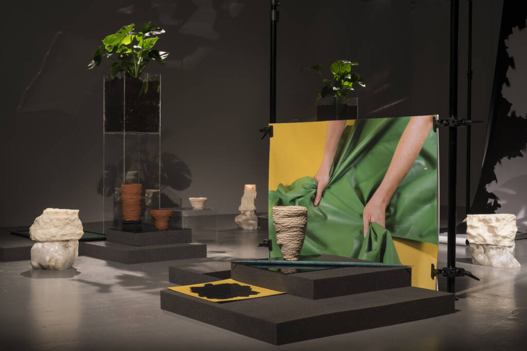 Gallery scene with stone sculptures, clay pots, plants in raised Perspex holders and a large central artwork held up by large legs on black steel rods showing hands stretching green material on a yellow background.