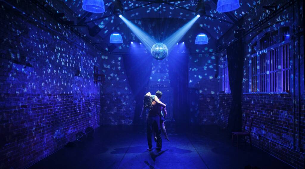 A pair of dancers stand in the centre of a room lit in blue and covered in the light of a disco ball. One dancer faces away from the viewer while lifting the second who lies back as their face is illuminated.