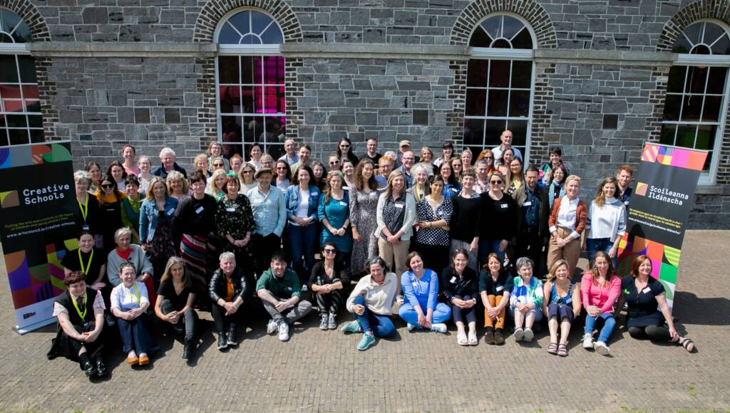 A large group of adults stand outside a stone building with large windows. They stand smiling between two large colourful Creative Schools pop-ups