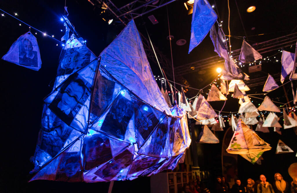 A series of decorative kite like sculptures hang from a series of wires with fairy lights.