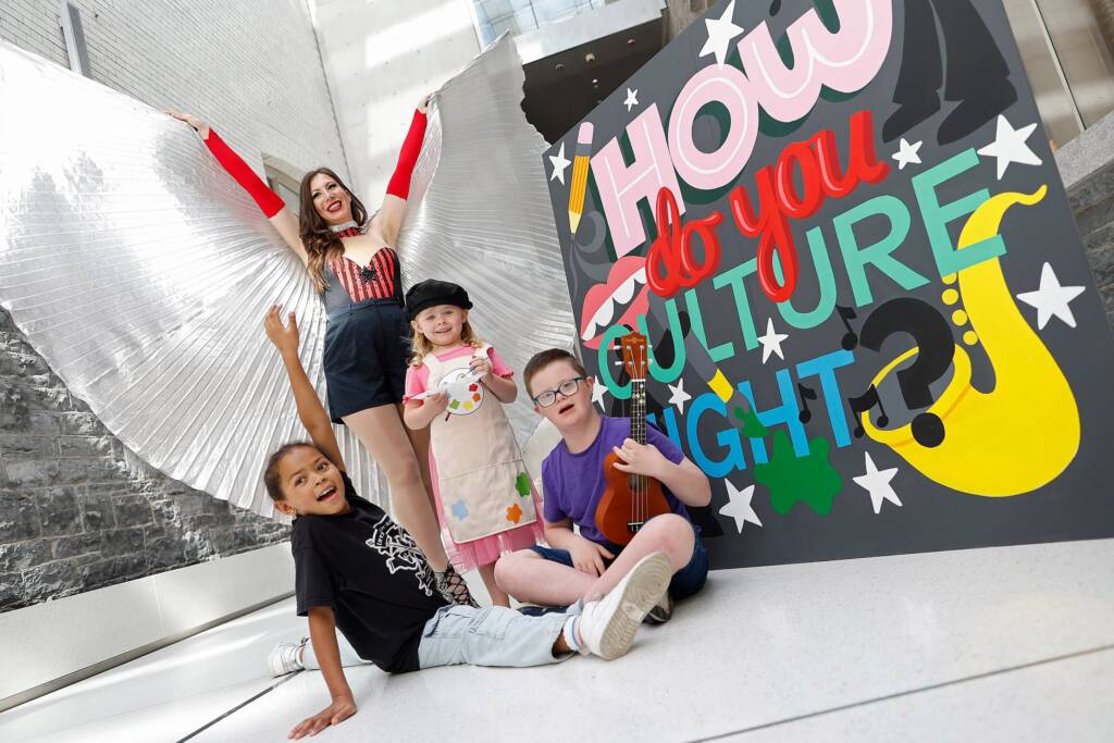 A group of young performers sit on the floor beside a colourful How Do You Culture Night sign and in front of a performer who again spreads their costume out to make wings