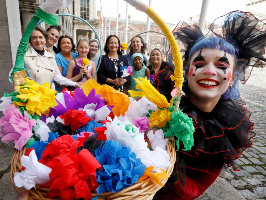 A group of people stand behind a bouquet of fake flowers in an outdoor setting. Some of the people hold fake flowers, while others hold hula hoops. They smile and look directly at the camera.