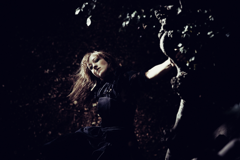 A performer is caught in motion, they wear a dark costume and look at the viewer from behind a knotted tree.