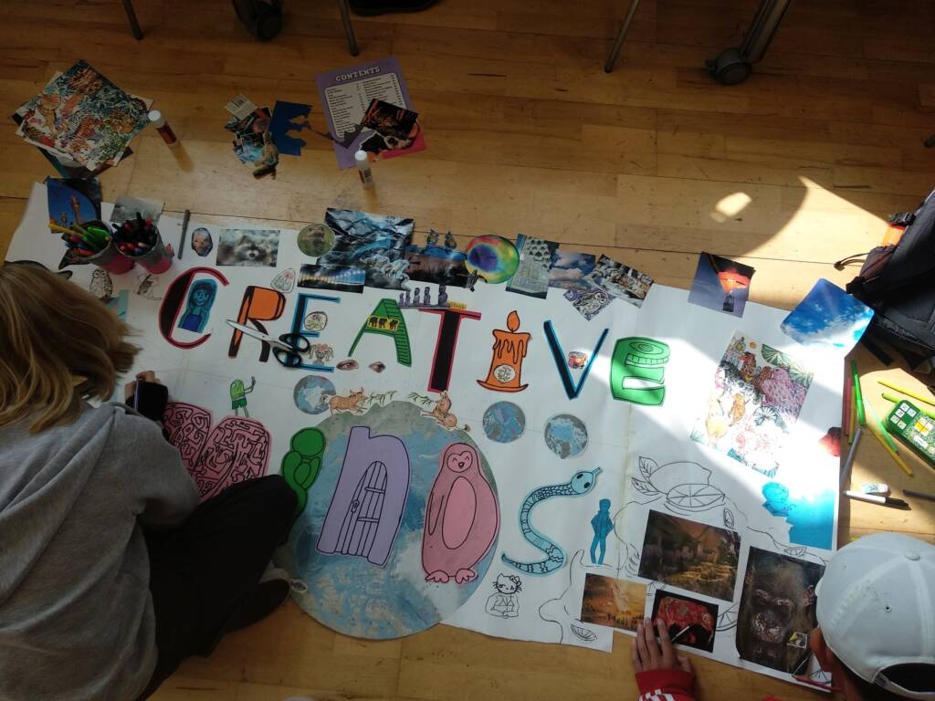 Students sit on the floor working on a colourful handmade poster collage spelling out 'CREATIVE MINDS' in different shapes and symbols