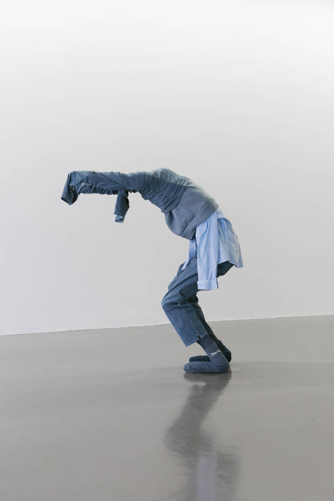 Statue of a figure covered in demin and other blue items of clothing including a blue shirt, socks and a jumper. The figure is bent over and head covered as they struggle to take off a denim shirt
