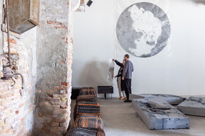 In search of Hy-Brasil Installation, two people hold up a piece of material in a gallery space. Around them are artworks on the floor and a partially plastered brick wall is in front of them