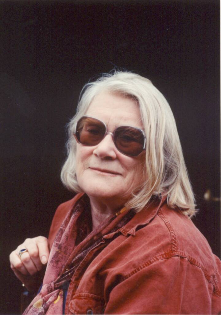 Headshot of writer Jennifer Johnston. She has shoulder length grey hair and wears brown sunglasses and a red jacket. She smiles and looks past the viewer.