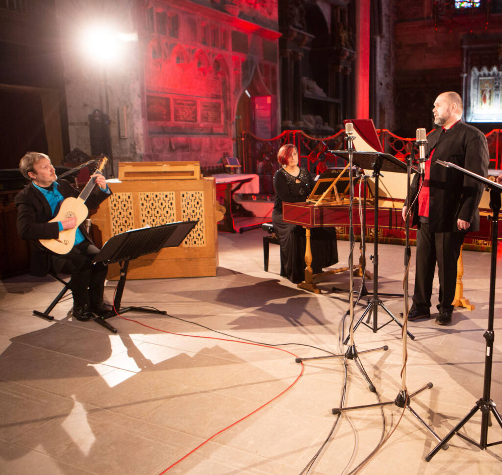 Three performers stand in a dramtically lit church interior. One plays a piano, one a guitar and one sings.