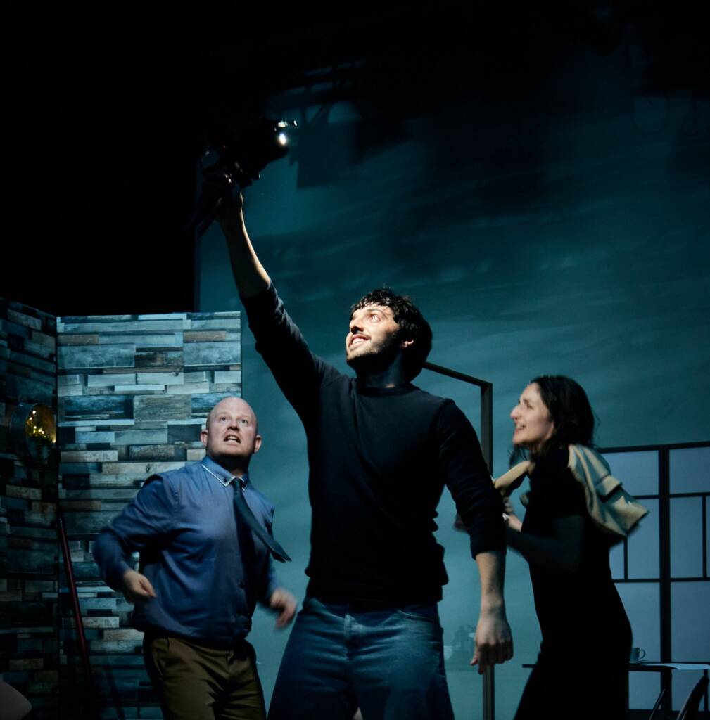 An actor on stage looks upwards towards the light with his hand raised. Behind him, two other actors cross the stage to walk towards each other.