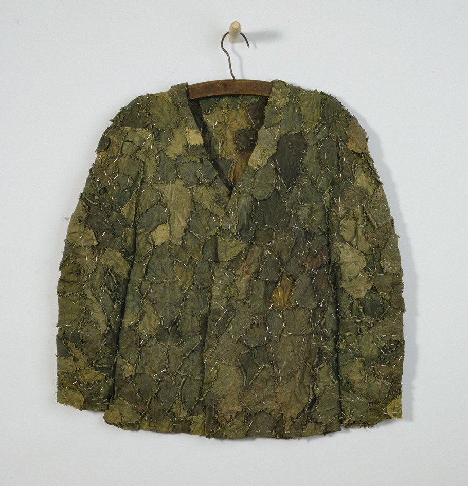 A coat made entirely from nettle leaves and pins hangs from a wooden hanger on a white wall