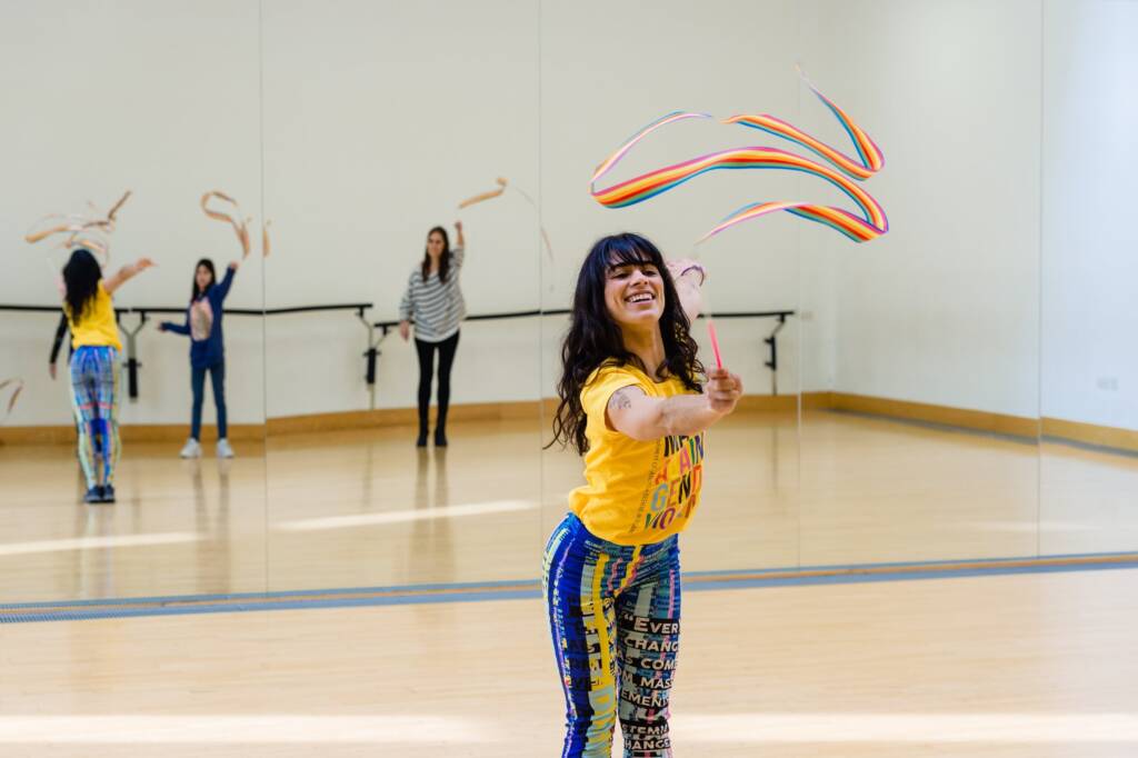 A teacher in bright clothing teaches young people ribbon dancing as seen reflected behind her in a wall of mirrors.