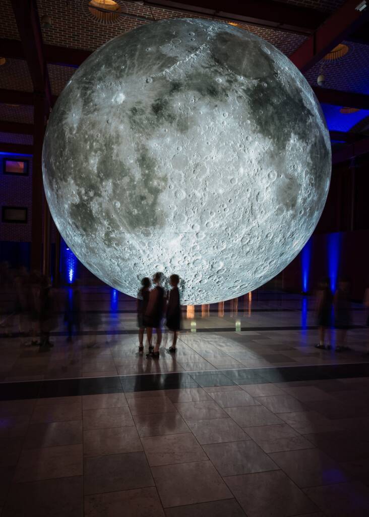 Three young people stand in front of a large moon sculpture light from within
