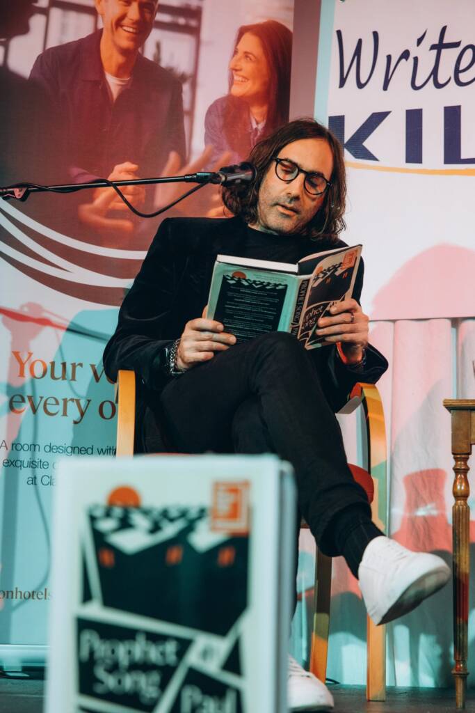 Author Paul Lynch sits in a chair at Write at the Sea. He reads from his book 'Prophet Song' while another copy of it sits in front, out of focus to the viewer.