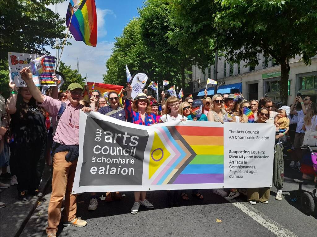 A group of Arts Council staff and friends hold a banner with the arts Council logo and inclusive pride flag