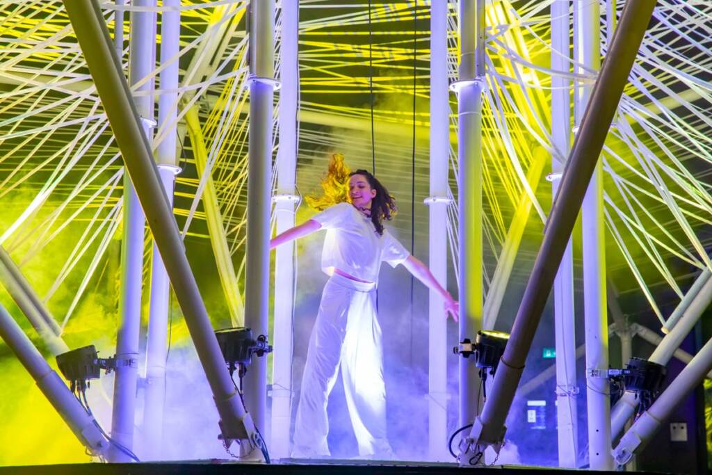 A performer dressed in white stands in the centre of a large structure light in white and yellow. The structure is composed of long white metal poles which are connected by a series or white ropes