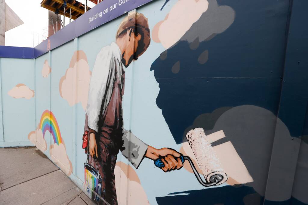 Mural on wooden fencing of a painter painting a blue sky with rainbows over a dark rainy stormy sky.