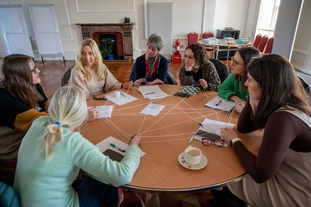 A group of women sit around a circular table. They all lean in in conversation as they hold a series of points of a star made from string