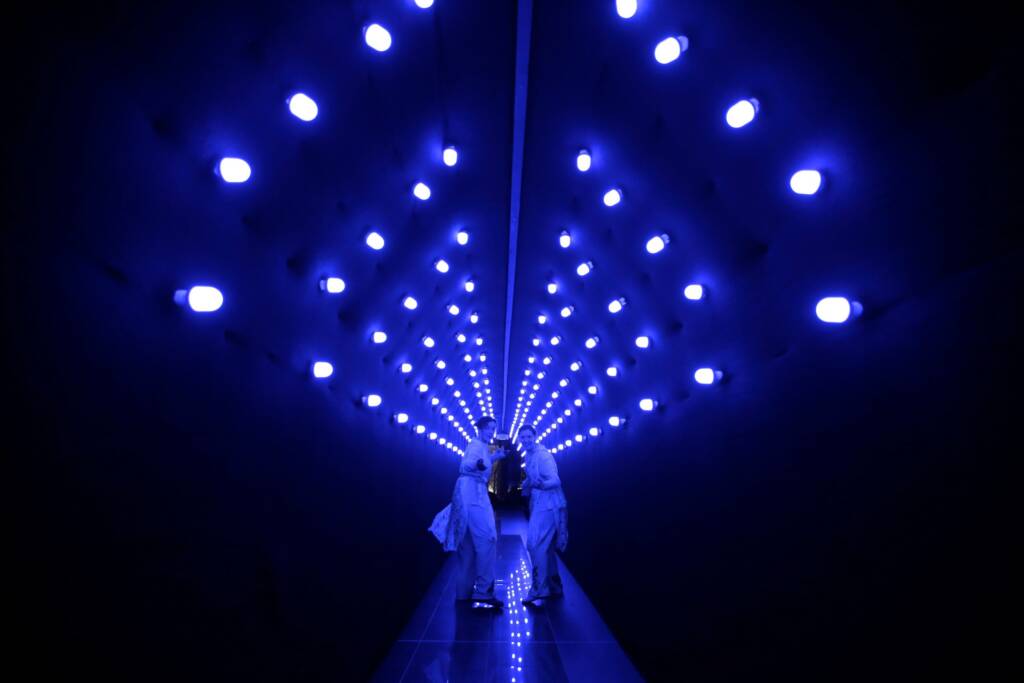 two performers turned back to look at the viewer in a hallway with blue lights above them