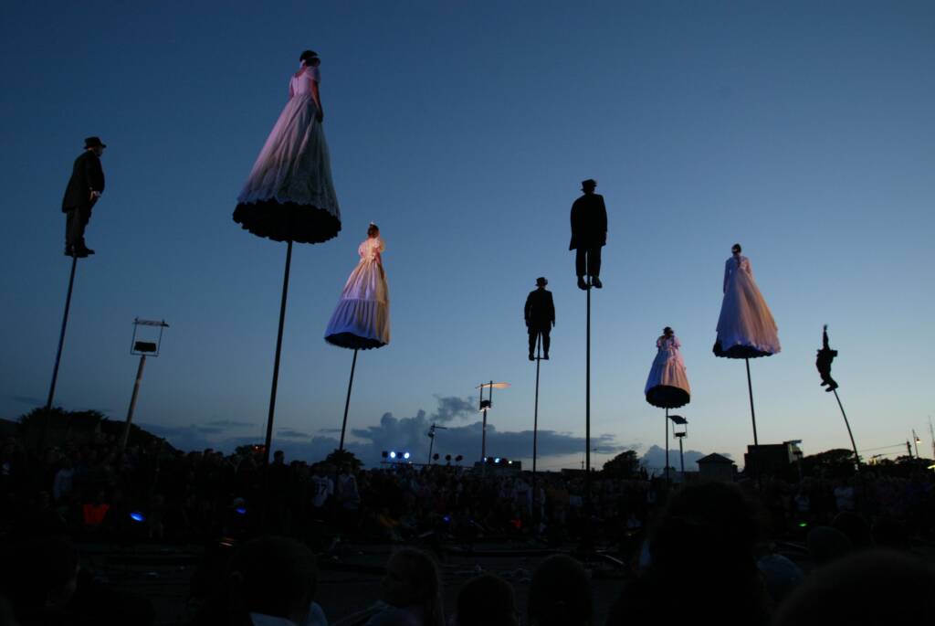Figures stand atop poles in victorian outfits above a crowd. They look away from the viewer in a dusk sky.