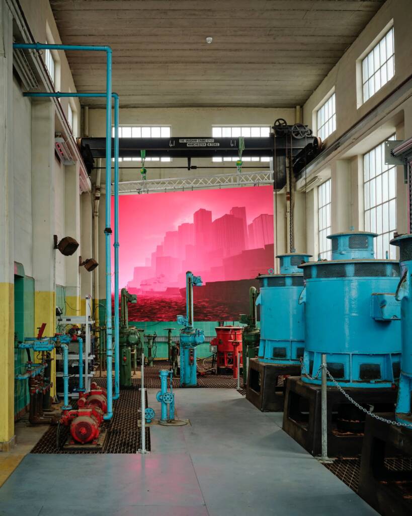 Large pieces of factory machinery in sky blue stand in front of a screen showing a vibrant pink cityscape.