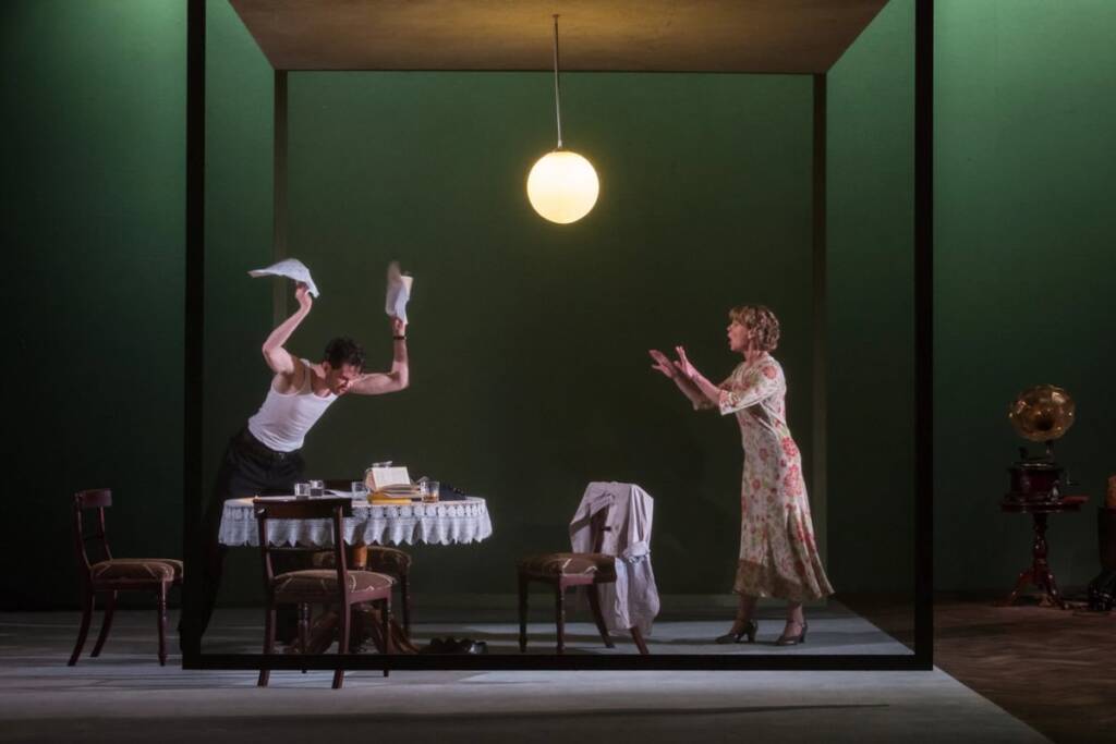 Scene from the Gate's production of The Glass Menagerie. A man wearing a white tank top is throwing pages aggressively at a table with a lace tablecloth while his partner raises her arms in a calming motion