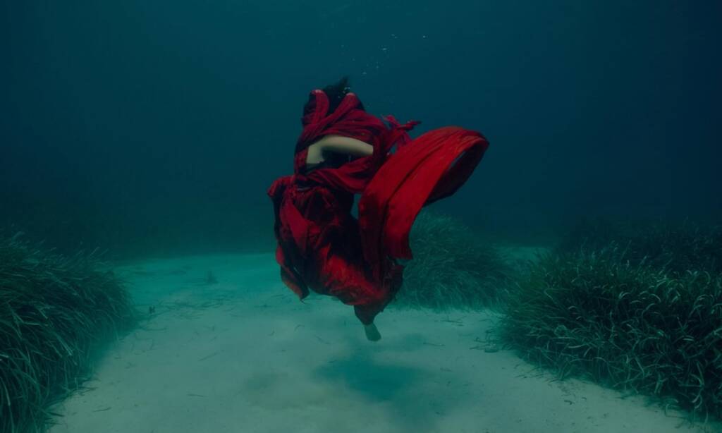 A dancer caught in motion underwater. They wear a red dress which floats up around their face and body and they appear to be at the ocean floor.