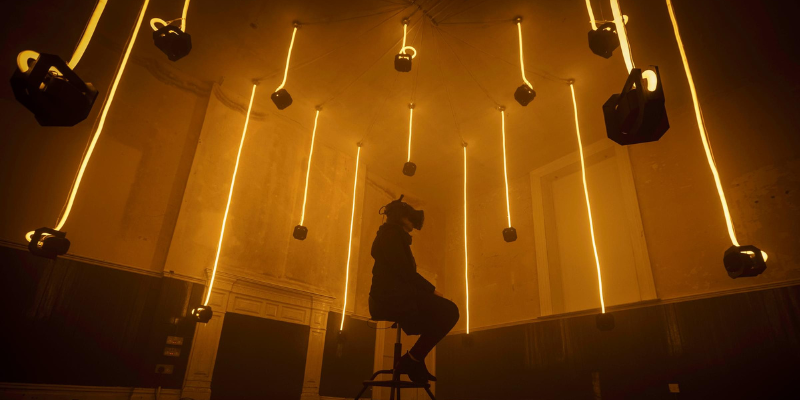 A person wearing a VR headset sitting in the centre of a room with hanging cameras on neon ropes.