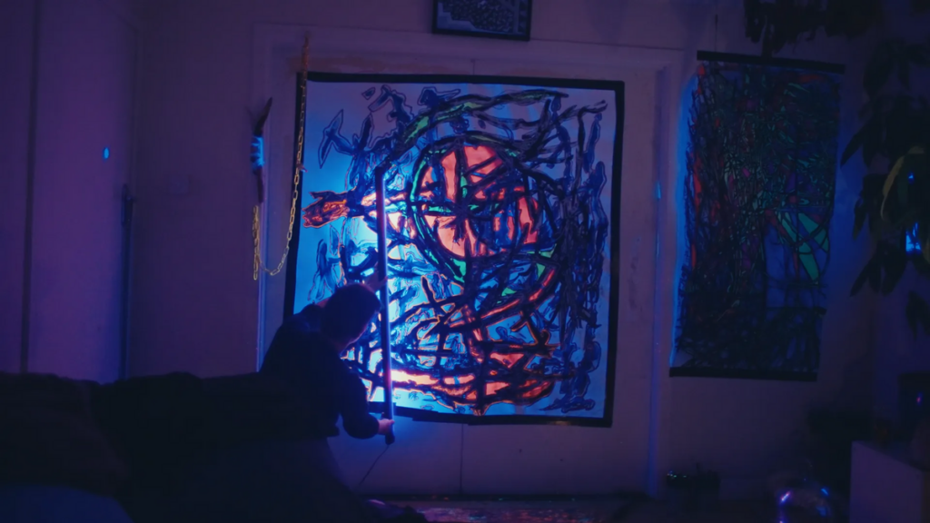 Still from Amanda. A person sits on the floor in a dimly lit room, gazing at a vibrant and abstract painting illuminated by colorful lights.